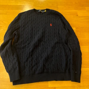 Svart stickad tröja från Polo Ralph Lauren - Säljer en klassisk svart stickad tröja från Polo Ralph Lauren med kabelstickat mönster. Tröjan har långa ärmar och en liten röd logotyp på bröstet. Perfekt för kyliga dagar!