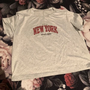 Grå t-shirt med New York-tryck - Säljer en grå t-shirt med rött 'New York' tryck och texten 'good night' under. T-shirten har en avslappnad passform och korta ärmar, perfekt för en casual look.