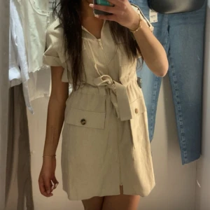 Beige kortklänning från Zara - Snygg beige kortklänning med dragkedja framtill och knytband i midjan från Zara