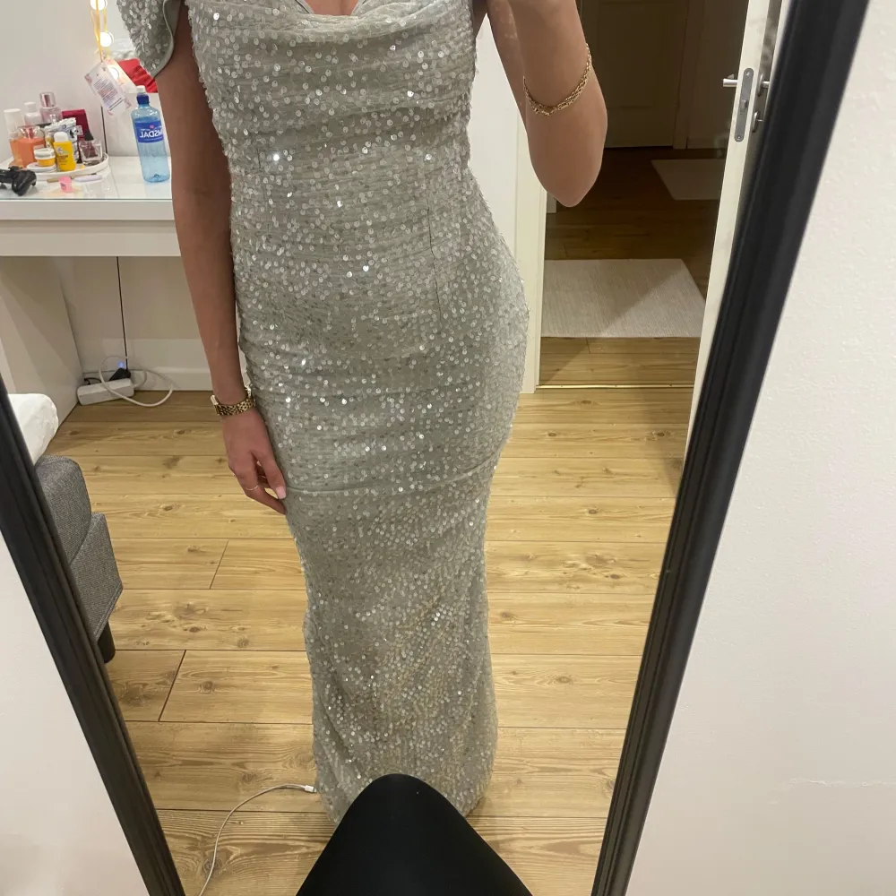 Snygg glittrig silverklänning med offshoulder-design och smala axelband. Klänningen är figurnära och har en elegant lång längd, perfekt för festliga tillfällen.. Mekot.