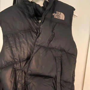 Svart dunväst från The North Face - Säljer en svart dunväst från The North Face med dragkedja. Perfekt för kyliga dagar och ger en sportig look. Västen har en hög krage och är idealisk för lager-på-lager-stil.