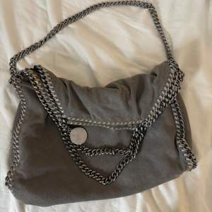 Säljer nu min fina grå axelväska från Stella McCartney Falabella i mellan storlek. Väskan har silvriga kedjedetaljer och en rymlig insida med rosa foder. Den är i fint skick och använd endast ett fåtal gånger. Ordinarie priset är ca 12.000 kr,  en av sidorna har ett minimalt hål som knappt syns vid användning (se sista bild), därav priset. Vid fler bilder eller mer äkthetsbevis är de bara att kontakta!