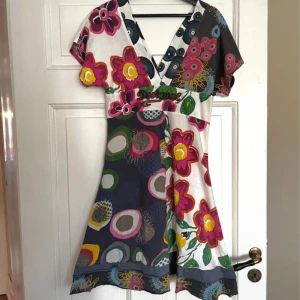 Färgglad blommig klänning - Säljer en unik och färgglad klänning med blommigt mönster i olika färger. Klänningen har en v-ringad hals och korta ärmar. Perfekt för att lysa upp garderoben!