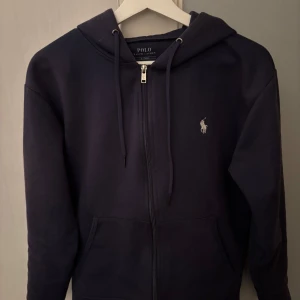 Mörkblå hoodie från Polo Ralph Lauren - Säljer en stilren mörkblå hoodie från Polo Ralph Lauren med dragkedja och det klassiska logotypbroderiet på bröstet. Säljer för den börjar bli lite för liten. Hoodien är i bra skick förutom att det tyvärr har blivit ett litet hål på insidan av tröjärmen. Men det är inget som syns i vanliga fall. Hör av dig vid frågor🙌