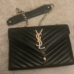 Svart quiltad axelväska från YSL - Snygg svart quiltad axelväska från YSL med guldfärgad kedja och logotyp. Väskan har en elegant design med ett rött foder inuti. 