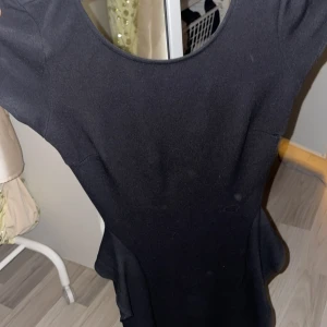 Svart klänning från Zara Trafaluc  - Elegant svart klänning från Zara Trafaluc med en figurnära passform och volangdetaljer i midjan. Klänningen har korta ärmar och en dragkedja i ryggen för enkel påklädning. Perfekt för en stilren look.