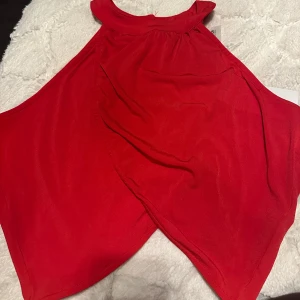 Röd ärmlös topp med omlott-design - Snygg röd ärmlös topp med omlott-design och hög hals. Perfekt för en kväll ute eller en speciell tillställning. Toppen har en elegant och modern stil. HELT NY OCH EJ ANVÄND.