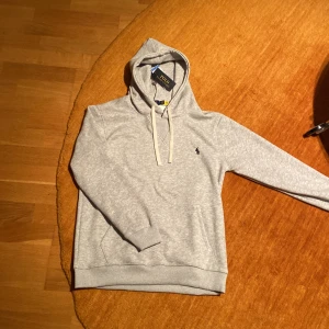 Grå hoodie från Polo Ralph Lauren - Säljer en stilren grå hoodie från Polo Ralph Lauren med det klassiska broderade logotypen på bröstet. Tröjan har långa ärmar och en bekväm passform med justerbar huva. Perfekt för en avslappnad look. Denna vara är sprillans ny och säljs pga att den inte passade på mig.