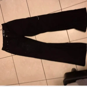 Svarta jeans med slitningar - Snygga svarta jeans med slitningar på benen. De har en klassisk design med fem fickor och knappgylf. Perfekta för en avslappnad stil.