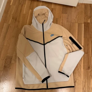 Beige och vit jacka från Nike - Säljer en snygg beige och vit Tech Fleece från Nike storlek L med dragkedja och huva. Jackan har svarta detaljer och en ficka på ärmen. Perfekt för en sportig look. Kan diskutera priset.