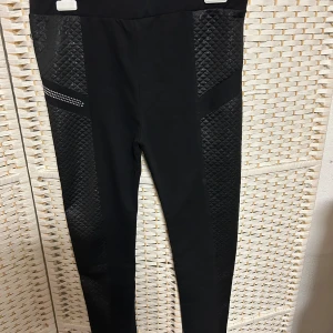 Svarta leggings med quiltade detaljer - Snygga svarta leggings med quiltade detaljer och glittriga accenter på sidorna. Perfekta för en stilren look med en twist. Högmidjade och tillverkade i ett stretchigt material för bästa komfort.