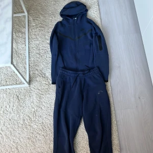 Mörkblå hoodie från Nike - Säljer en mörkblå Nike tech. Bra skick på både tröjan och byxorna, tröjan=158-170cm byxorna=158-170cm. Priset är inte hugget i en sten, hör av er vid frågor. 