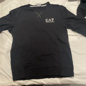 Svart tröja från Emporio Armani - Snygg svart tröja från Emporio Armani med EA7-logga på bröstet. Tröjan har en rund halsringning med dekorativt kryssmönster. Perfekt för en stilren look.