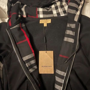 Svart hoodie från Burberry - Snygg svart hoodie från Burberry med klassiskt rutigt mönster i huvan. Den har dragkedja framtill och en justerbar huva med snörning. Perfekt för en stilren look. 