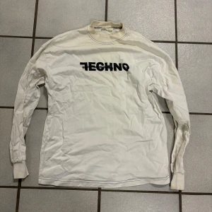 Vit långärmad tröja från ALYX - Säljer en vit långärmad tröja från ALYX med texten 'TECHNO' i svart på framsidan. Tröjan har en ribbad krage och texten 'ALYX/NEW HAPPINESS' på baksidan vid nederkanten. Perfekt för en avslappnad stil. Köpt i Japan 