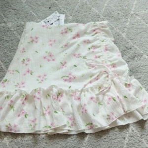 Blommig kjol  - Charmig vit kjol med rosa blommönster från Gina Tricot. Kjolen har volangdetaljer och en omlott-design som ger en romantisk känsla. Perfekt för somriga dagar. Med prislapp kvar och aldrig använd. Storlek S.