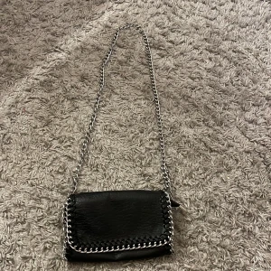 Svart axelväska från Tiamo - Snygg svart axelväska från Tiamo med kedjedetaljer. Väskan är tillverkad i syntetmaterial och har en praktisk dragkedja. Perfekt för att ge din outfit en edgy touch. Har en liten fläck på insidan går säkert att tvätta bort💗