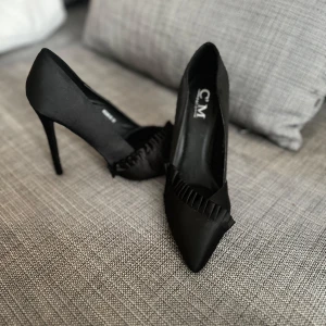 Svarta klackar  - Eleganta svarta klackar med spetsig tå och hög klack. Skorna har en dekorativ detalj framtill som ger en extra touch av stil. Perfekta för en kväll ute eller en speciell tillställning. Klackarna är aldrig använda