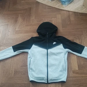 Nike tech - Säljer en snygg grå och svart hoodie från Nike med dragkedja och huva.  Säljer den på grund av att jag tycker den är för stor och vill nu blir av med den den är hel ny aldrig andvänd