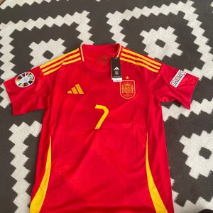 Röd Spanien fotbollströja med nummer 7 - Snygg röd Spanien fotbollströja från Adidas med gula detaljer och nummer 7 på framsidan. Tröjan har korta ärmar och spelarens namn 'Morata' på baksidan. Perfekt för fotbollsfans!