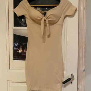 Beige klänning med knyt - Trendig beige klänning med knyt framtill och offshoulder-design. Den har en tight passform och är tillverkad i ett ribbat material som ger en snygg siluett.