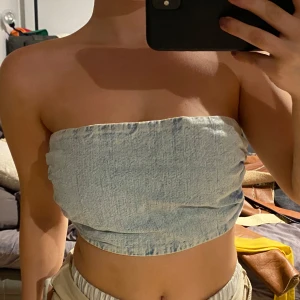 Ljusblå jeans tubtopp - Trendig ljusblå tubtopp i jeansmaterial med smockdetaljer på sidorna för en bekväm passform. Perfekt för sommardagar och festivaler.