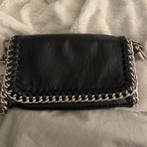 Svart axelväska från Tiamo - Snygg svart axelväska från Tiamo med silverkedja och dragkedja. Väskan har en cool design med flätade detaljer längs kanterna och en avtagbar kedjerem. Perfekt för att ge din outfit en edgy touch. Den har en defekt man ser på sista bilen❤️