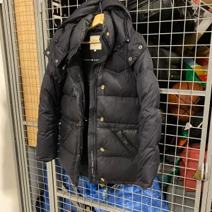 Svart dunjacka med huva - Snygg svart dunjacka från abonnerande med huva och dragkedja. Jackan har knappar och praktiska fickor framtill. Perfekt för kyliga dagar. 5000kr för en ny, säljer min för 1000kr, finns få skråmor som ja visar på bilden, säg till om ni vill ha fler bilder!