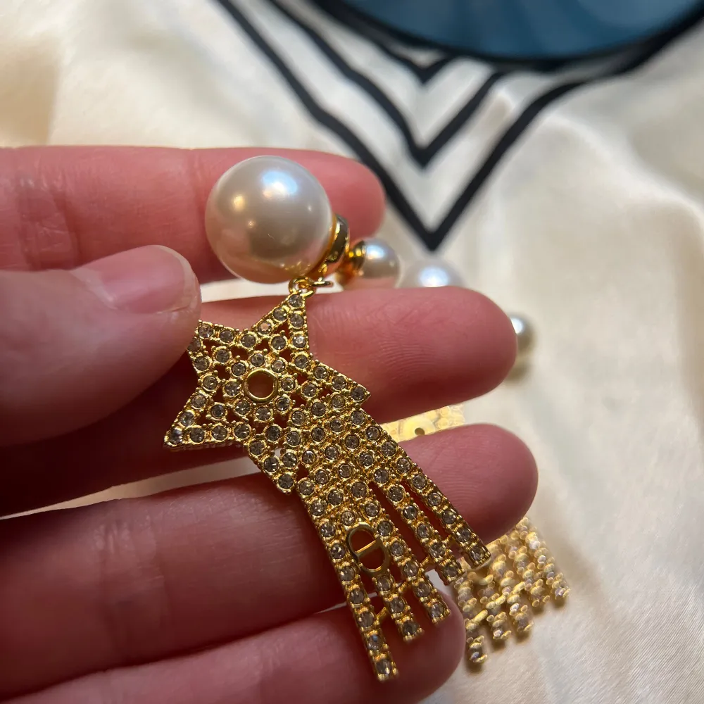 Snygga örhängen i guld med stjärnformade hängen dekorerade med glittrande stenar. Örhängena har stora vita pärlor som fäste och mindre pärlor ovanför stjärnorna. Perfekta för att ge en glamorös touch till din outfit.. Asusteet.