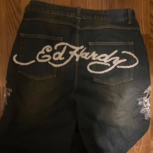 Baggy jeans från Ed Hardy x Urban Outfitters - Coola mörkbruna jeans från Ed Hardy (köpt på urban); unikt broderad och med logotyp på bakfickorna. OBS!! Väldigt slitna vid bensluten (annars inga defekter) + jag klippte dem kortare vid köp för att passa min längd bättre. Ny längd: ca 107cm. Midja: 46cm rakt över.