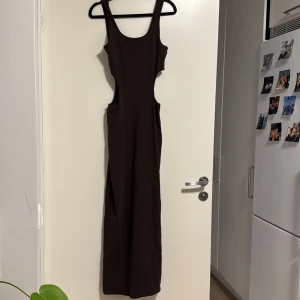 Brun långklänning från Weekday - Snygg brun långklänning från Weekday med ärmlös design och djupa utskärningar i sidorna. Perfekt för en stilren och modern look. Tillverkad i ett mjukt och bekvämt material.