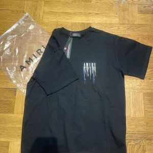 Svart t-shirt från Amiri - Snygg svart t-shirt från Amiri med ett coolt tryck av droppande färg i vitt, blått och guld. Perfekt för en stilren look med en twist. T-shirten är kortärmad och har en normal passform.