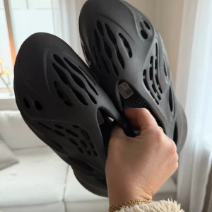 Yeezy Foam Shoes - Säljer ett par svarta Yeezy Foam med unik och futuristisk design. Skorna har en luftig struktur med flera öppningar och en robust sula. Perfekta för en modern och stilren look. Använda endast två gånger och är i nyskick. Behövs bara torkas av då dem samlat på sig damm. Stl 40.5 