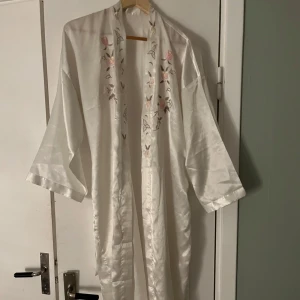 Vit kimono med broderade blommor - Elegant vit kimono med vackra broderade blommor i rosa och beige. Den har ett knytband i midjan. Perfekt för en avslappnad och stilfull look.