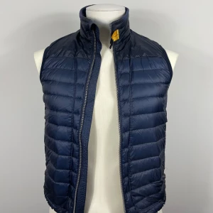 Mörkblå dunväst från Parajumpers - Snygg mörkblå dunväst från Parajumpers med dragkedja och hög krage. Västen har en broderad logga på ryggen och en gul detalj vid halsen. Perfekt för kyliga dagar. Köpt från parajumpers hemsida.