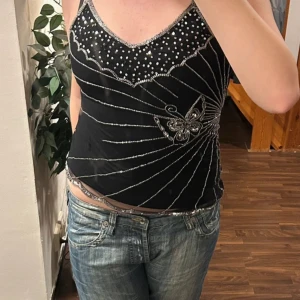 Svart linne med silverdetaljer - Snyggt svart linne med glittriga silverdetaljer och en fjärilsdesign. Toppen har en v-ringad hals och är ärmlös, perfekt för en festlig kväll. Passar bra med jeans eller kjol för en stilren look.