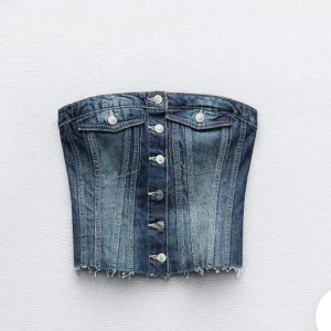 Jeans korsett med knappar - Trendig jeans korsett med knappar framtill och rå kant. Perfekt för en edgy look. Den har en klassisk denimfärg och stilrena sömmar.