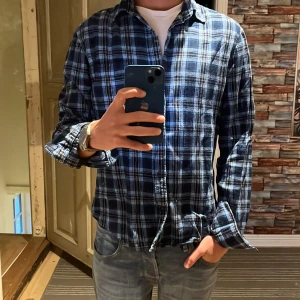 Blå och svart rutig skjorta från Dressmann - Säljer en snygg blå och svart rutig skjorta från Dressmann i storlek S. Skjortan har långa ärmar och en klassisk krage, perfekt för både vardag och fest. Den är i mycket bra skick och har en bröstficka för extra stil. Passar perfekt till jeans för en avslappnad look.