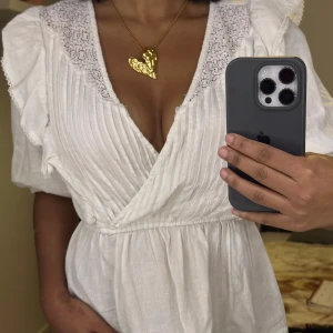 Vit volangtopp med spetsdetaljer - Superfin vit volangtopp med spetsdetaljer vid axlarna och volanger längs ärmarna. Den har en v-ringad design och trekvartsärmar, perfekt för sommar och vår. Toppen är i ett lätt och luftigt material, vilket gör den idealisk för varma dagar. 