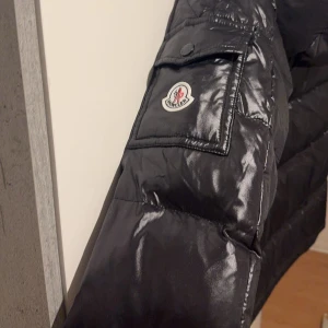 Svart dunjacka från Moncler - Säljer en snygg svart dunjacka från Moncler i mycket bra skick. Jackan har en glansig finish och är quiltad för extra värme. Den har en dragkedja framtill och praktiska fickor med knappar. Perfekt för kalla vinterdagar!                  Jackan är helt oanvänd, då den var lite för stor, skulle säga den passar M-L Perfekt. 