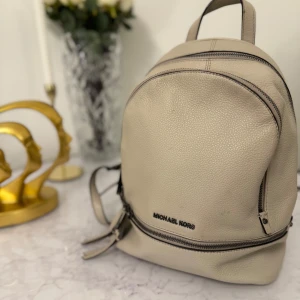 Beige ryggsäck från Michael Kors - Snygg och stilren beige ryggsäck från Michael Kors i mycket bra skick. Den har en praktisk dragkedja och är perfekt för både vardag och utflykter. Ryggsäcken är gjord i skinn och har en klassisk design som passar till alla tillfällen.