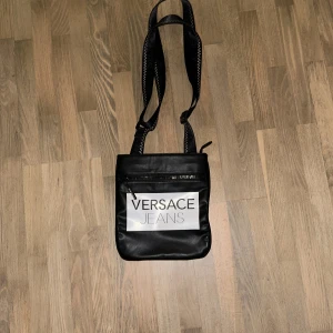 Svart axelväska från Versace Jeans - Snygg svart axelväska från Versace Jeans i syntetmaterial. Väskan har en justerbar axelrem och ett stort fack med dragkedja. Perfekt för vardagsbruk eller en kväll ute. Den har en stilren design med märkets logga tydligt på framsidan.