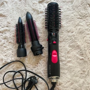 Remington varmluftsbortse - Bild på en Remington varmluftsborste med tre utbytbara borsthuvuden. Den har en svart och rosa design med en sladd för strömförsörjning. Perfekt för att styla håret med volym och lockar.