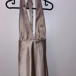 Champagnefärgad halterneckklänning - Elegant champagnefärgad halterneckklänning i satin. Perfekt för festliga tillfällen med sin mjuka glans och stilrena design. Klänningen har en vacker passform och knytning i nacken för en sofistikerad look.