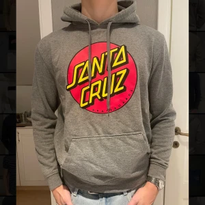 Grå hoodie från Santa Cruz - Säljer en grå hoodie från Santa Cruz med deras ikoniska logga i rött och gult på framsidan. Den har en stor ficka fram och justerbar huva med snören. Perfekt för en avslappnad stil. Bra skick!