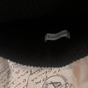 Svart stickad mössa från Moncler - Säljer en snygg svart stickad mössa från Moncler. Den har en klassisk ribbad design och ett broderat Moncler-märke framtill. Perfekt för att hålla sig varm och stilren under kalla dagar. Passar till alla vinteroutfits!