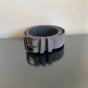 Grått mockabälte - Säljer ett stilrent grått bälte i mocka. Perfekt för både vardag och fest. Bältet har en klassisk design med en robust spänne i metall. Passar till de flesta outfits och ger en snygg touch till din stil. Ett måste i garderoben!. Skick 10/10 aldrig använt då det inte kändes min stil när jag fick hem den 