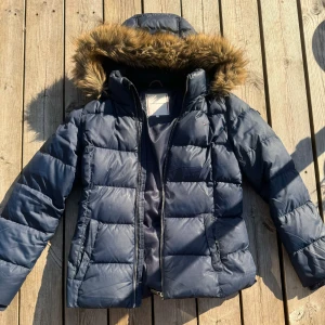 Blå dunjacka från Tommy Hilfiger - Säljer en snygg blå dunjacka från Tommy Hilfiger. Jackan har en mysig pälskantad huva och dragkedja framtill. Perfekt för kalla vinterdagar! Den är i  bra skick, förutom en liten lagning på ena axeln (se sista bilden) och passar både till vardags och lite finare tillfällen. Storlek 176 (motsvarar S)