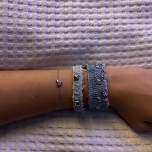 Armband i jeans och silver - Två coola armband, ett i jeans med nitar och ett tunt silverarmband med ett hjärta. Perfekt för att ge din outfit en edgy touch. Jeansarmbandet har en rå kant och nitar som ger en rockig känsla, medan det tunna silverarmbandet är mer subtilt och elegant.