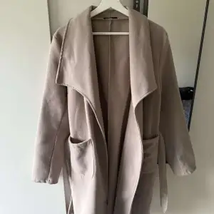 Säljer en snygg beige kappa från Boohoo i mycket bra skick. Den har långa ärmar och ett bälte i midjan för en elegant look. Perfekt för höst och vår, och den är supermjuk och bekväm. Passar både till vardags och fest!